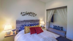 A bedroom with a wooden headboard, a bed with white bedding, two bedside lamps, a decorative wall piece, and a window with sheer curtains.