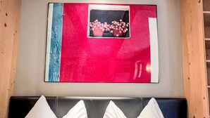Een kamer met een bed, een zwart hoofdbord en twee witte kussens. Daarboven een kunstwerk met een rode achtergrond, en een raam met roze bloemen.