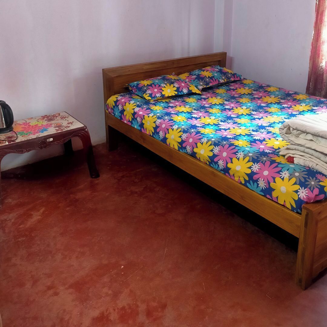 Un lit simple avec une couvre-lit à motifs floraux colorés, une tête de lit en bois et une petite table en bois recouverte d’une nappe à motifs floraux.
