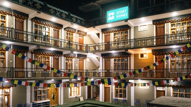 A multi-story hotel with a sign reading "Story Homes." It features balconies with railings, multiple windows, and a courtyard with chairs. - Story Homes (Kaza)