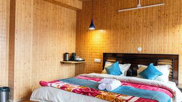 A wooden-paneled room with a bed, a hanging light fixture, a shelf with a kettle and cups, and a trash bin.