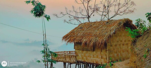 A traditional bamboo house with a thatched roof, elevated on stilts, surrounded by bamboo and trees. - Mirinja Valley Megh Machang Resort (Lama)