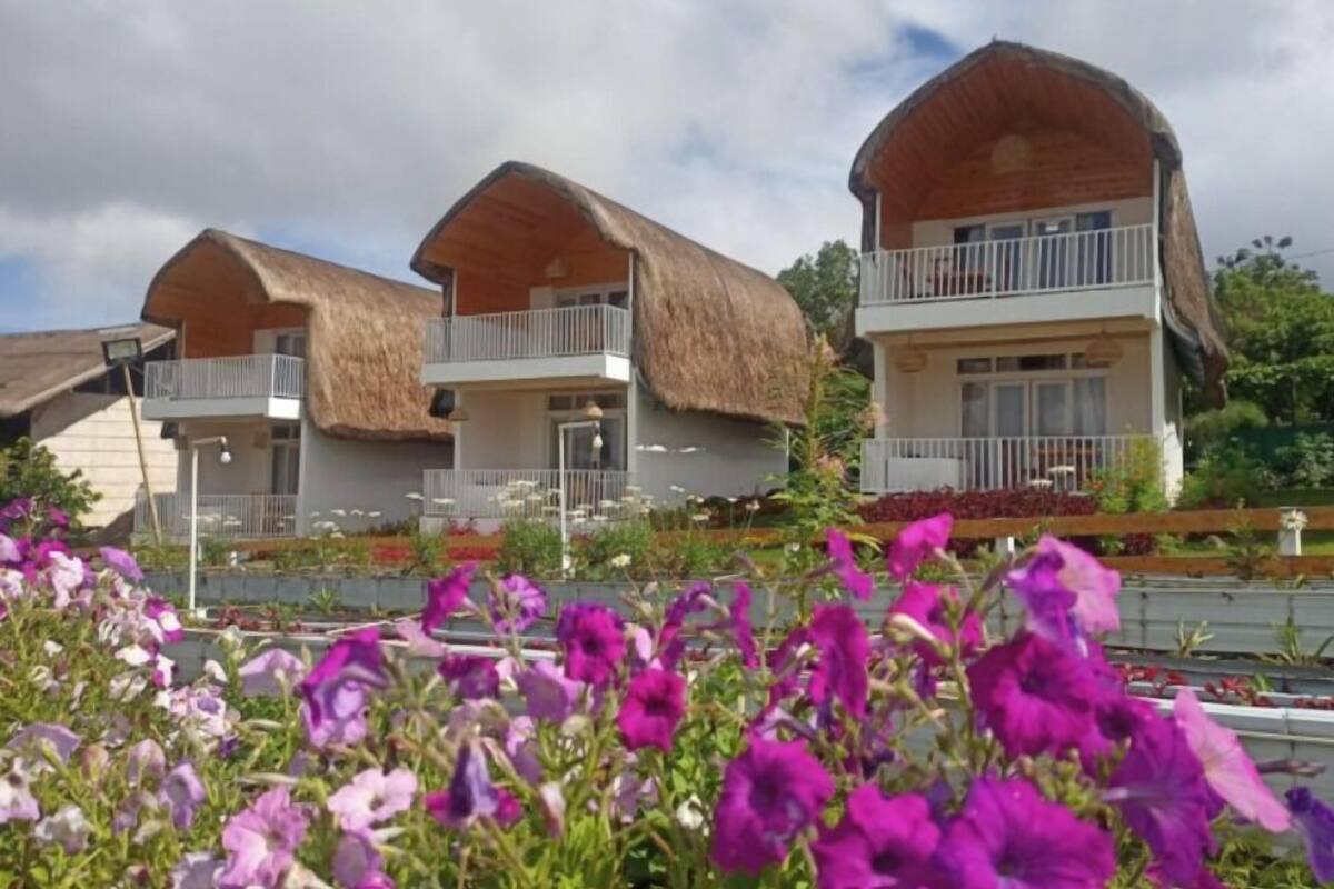 Una hilera de cabañas modernas de dos pisos con techos de paja y balcones, rodeadas de coloridos jardines florales.