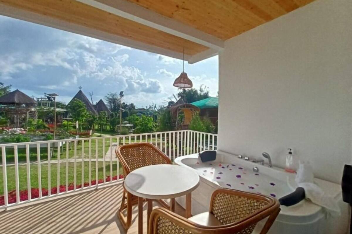 Un balcón con una mesa y sillas, una lámpara colgante, y vistas a un jardín y edificios al fondo.