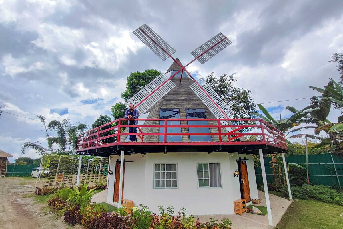 Una casa con un molino de viento en el techo, un balcón con barandillas rojas y un pequeño jardín delantero.