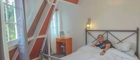 Un dormitorio con estructura metálica para la cama, techo de madera con vigas a la vista, una mesita de noche pequeña y una ventana con cortinas.
