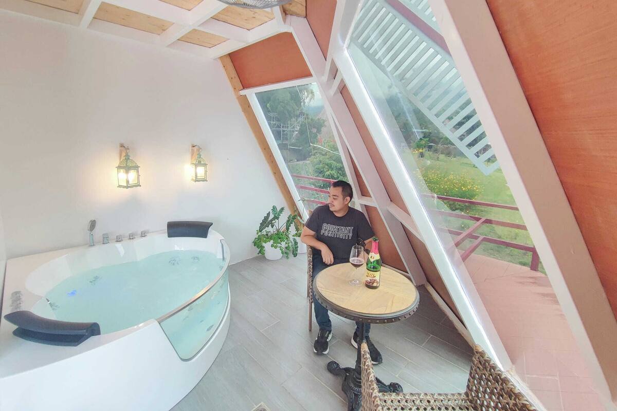 Una habitación con una bañera grande, una mesita redonda con una botella y un vaso, y vistas a espacios verdes al exterior.