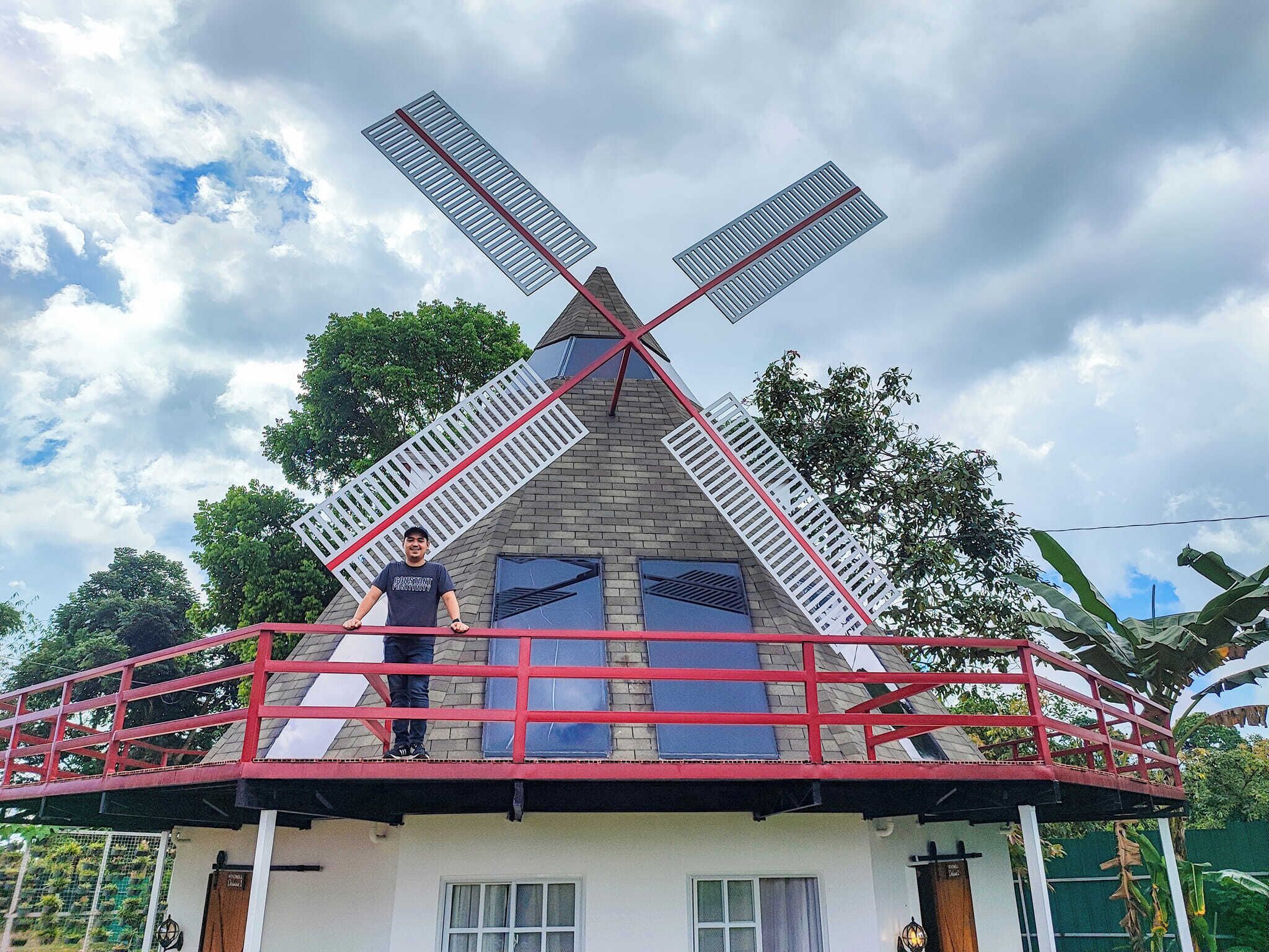 Een huis met een windmolenconstructie op het dak, een balkon met een rode reling, en een persoon die op het balkon staat.