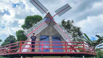 Een huis met een windmolenconstructie op het dak, een balkon met een rode reling, en een persoon die op het balkon staat.