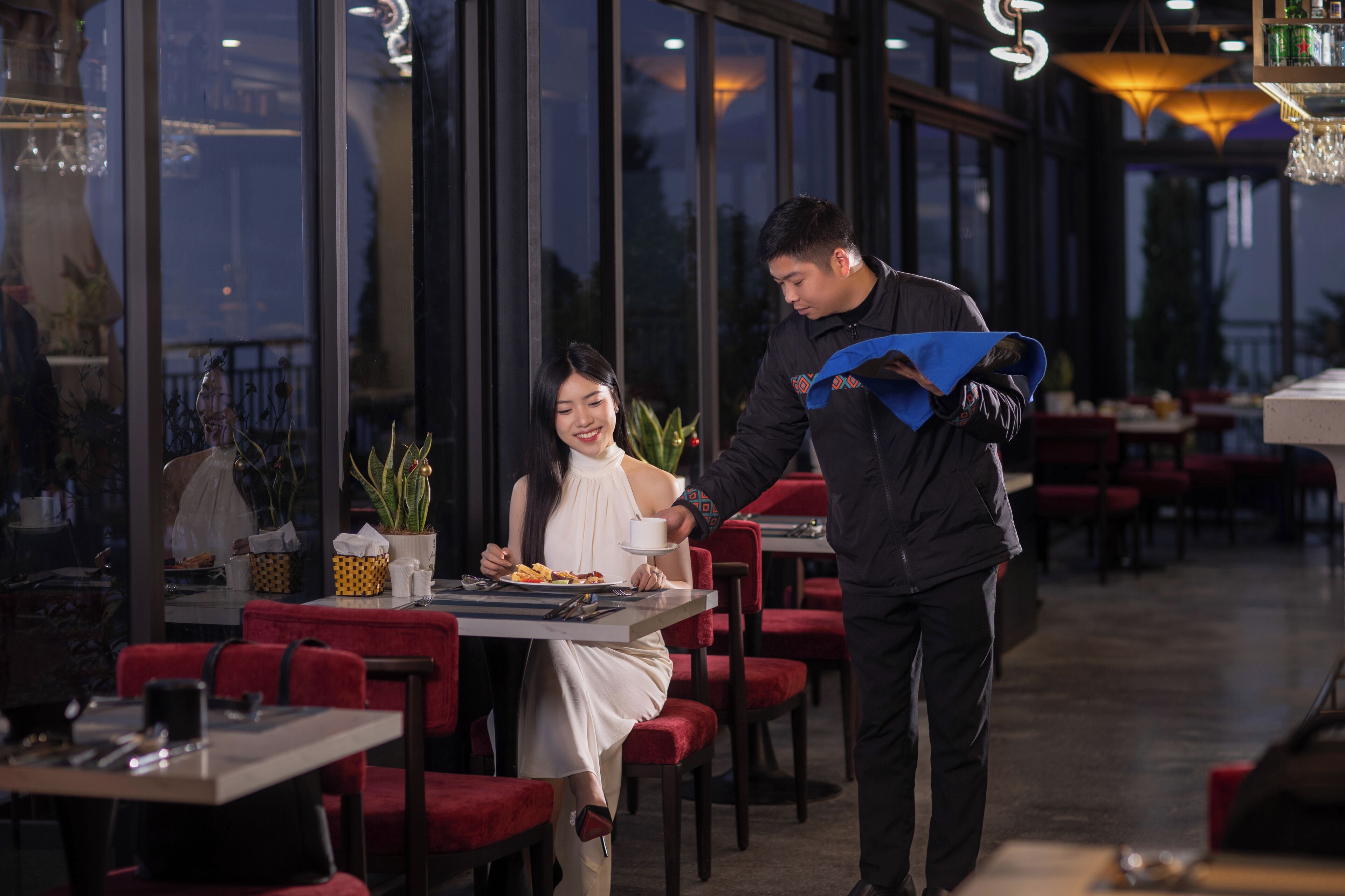 A restaurant with red upholstered chairs and tables, large windows, and a waiter serving a customer.
