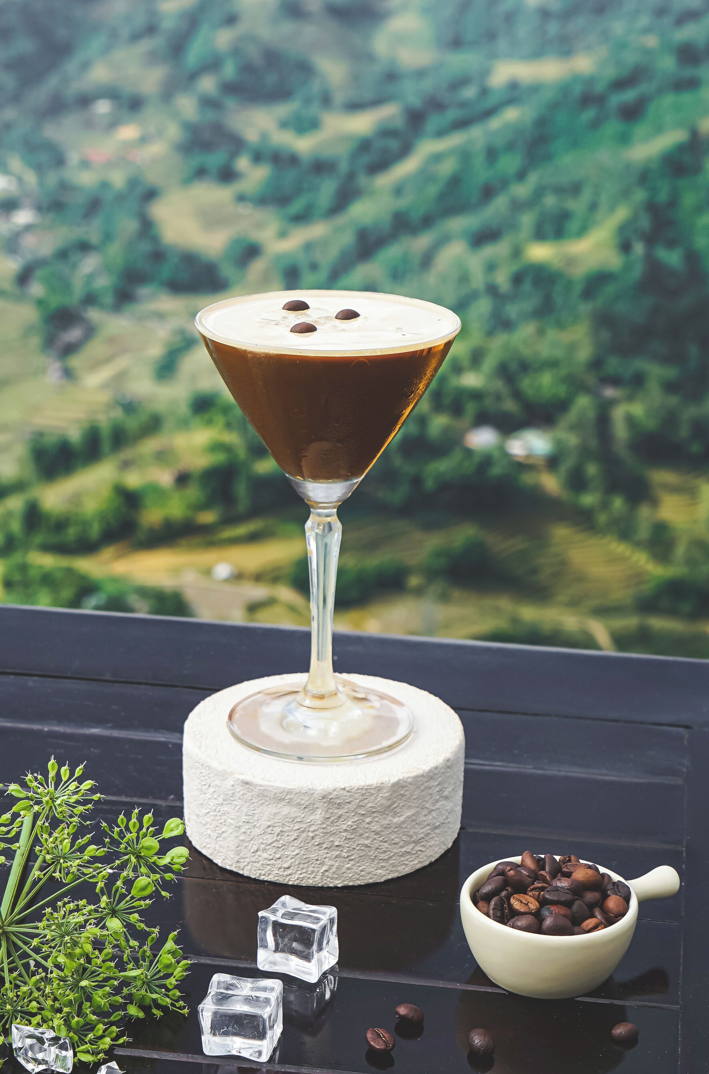 A martini glass with a frothy top and chocolate shavings, a small bowl of coffee beans, and ice cubes on a reflective surface.
