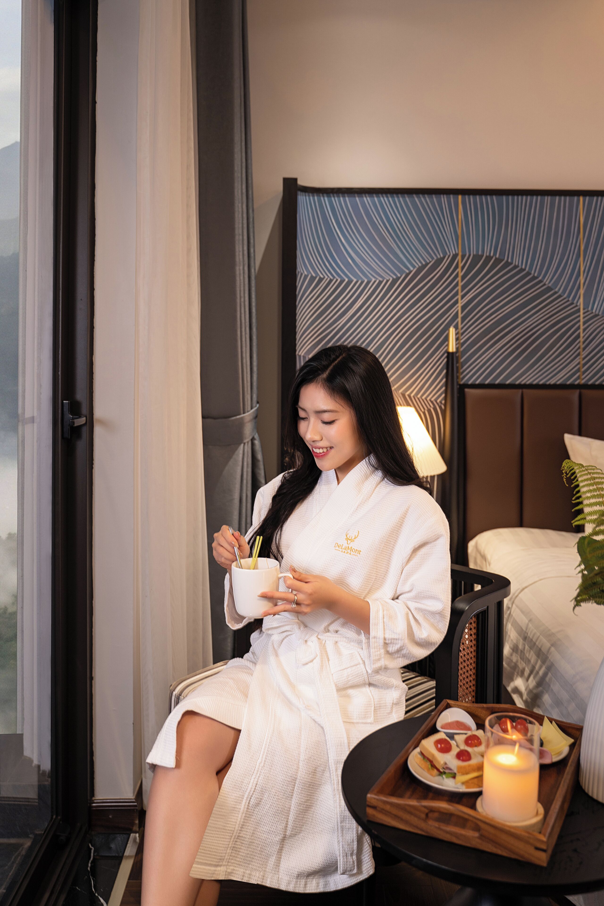 A woman in a white robe sitting by a window with a tray of food and a candle.