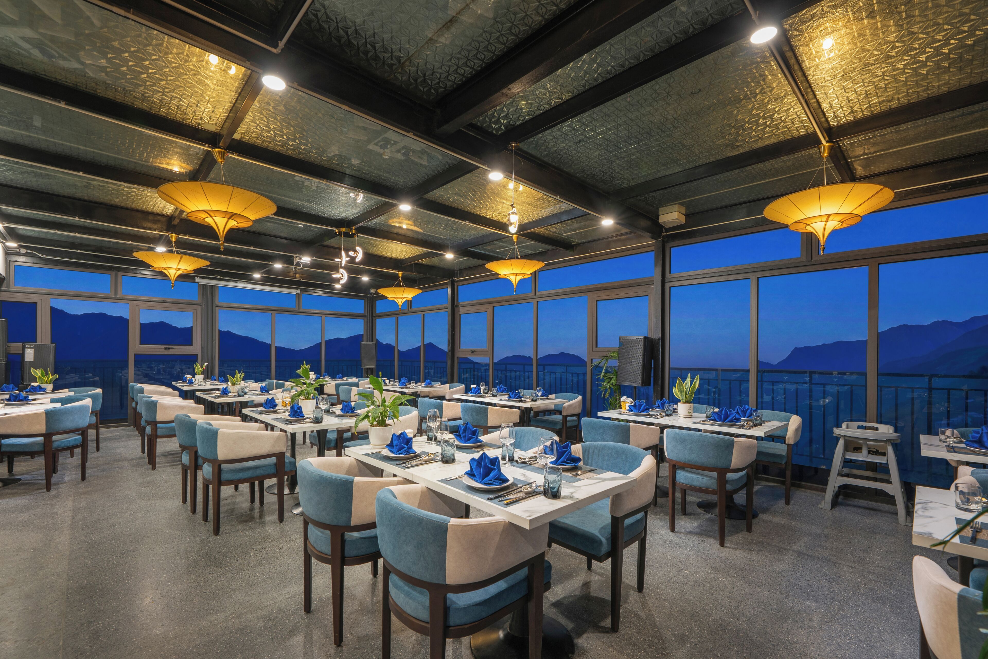 A restaurant with multiple tables and chairs, large windows, and hanging lamps.