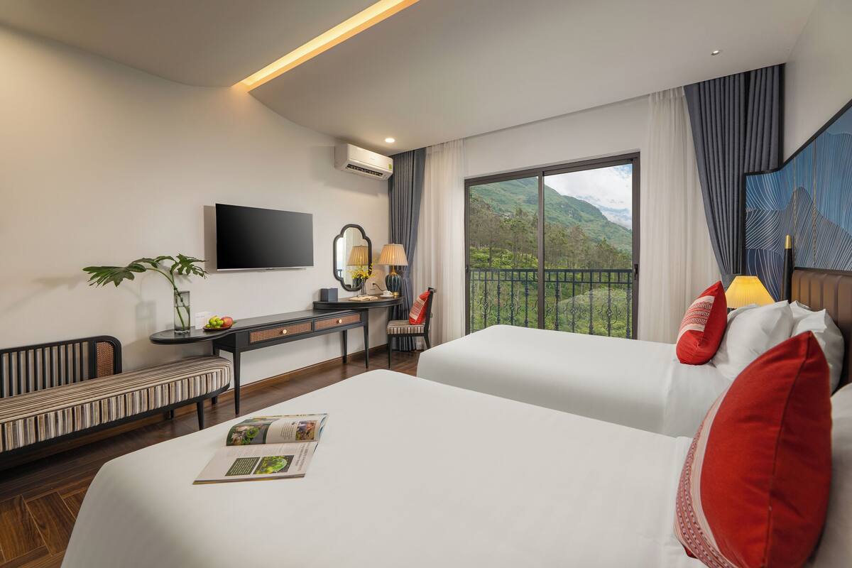 A hotel room with two beds, a desk, a TV, and a balcony with a mountain view.