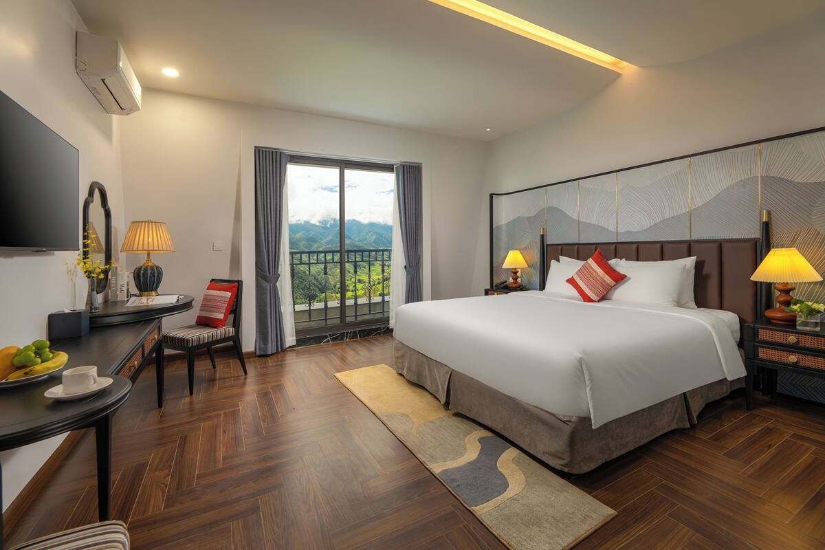 A hotel room with a large bed, a desk with a chair, a TV, and a view of mountains through a balcony door.