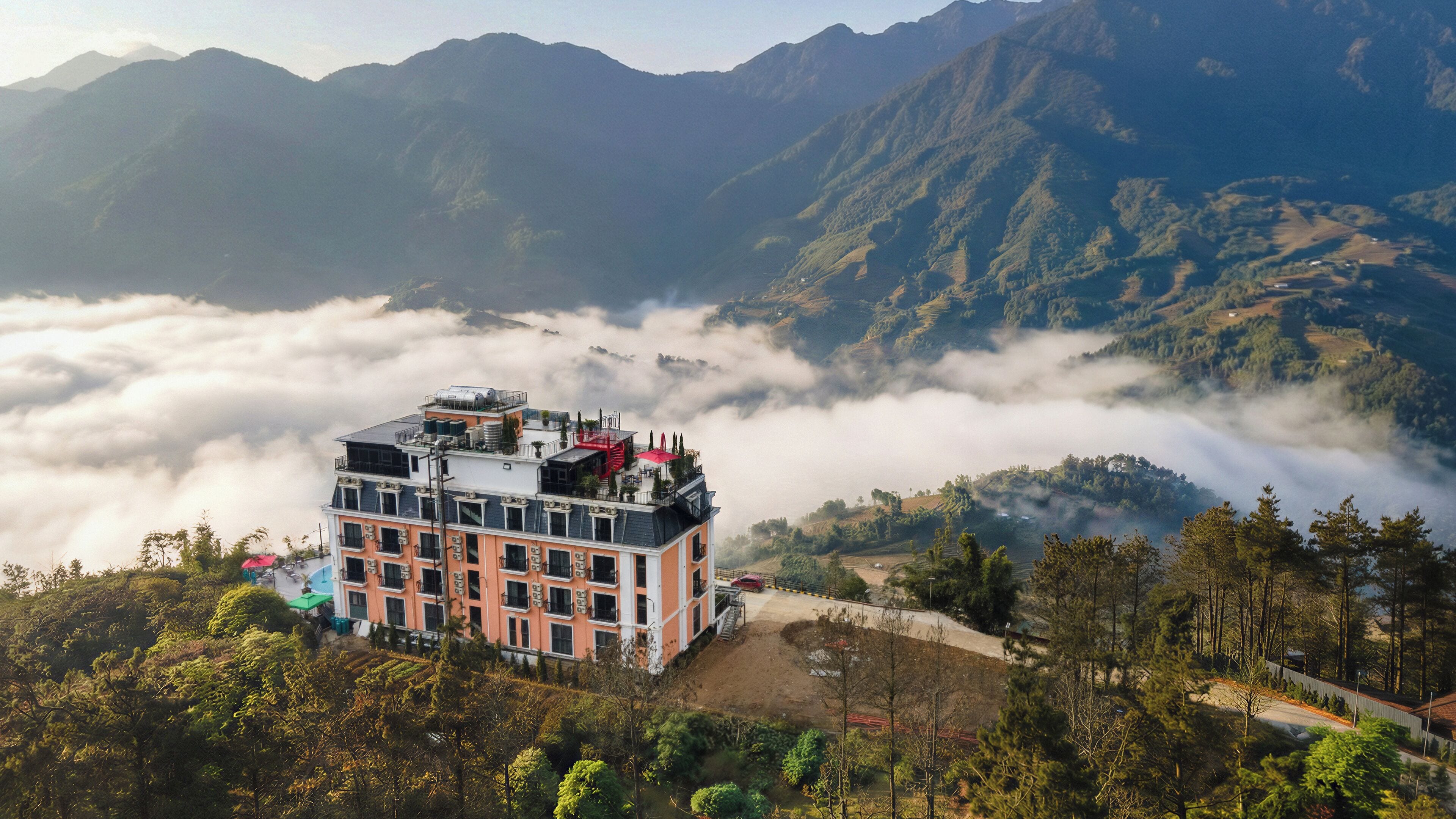 Hotel de vários andares com terraço no telhado, rodeado por colinas arborizadas e um vale de nuvens.
