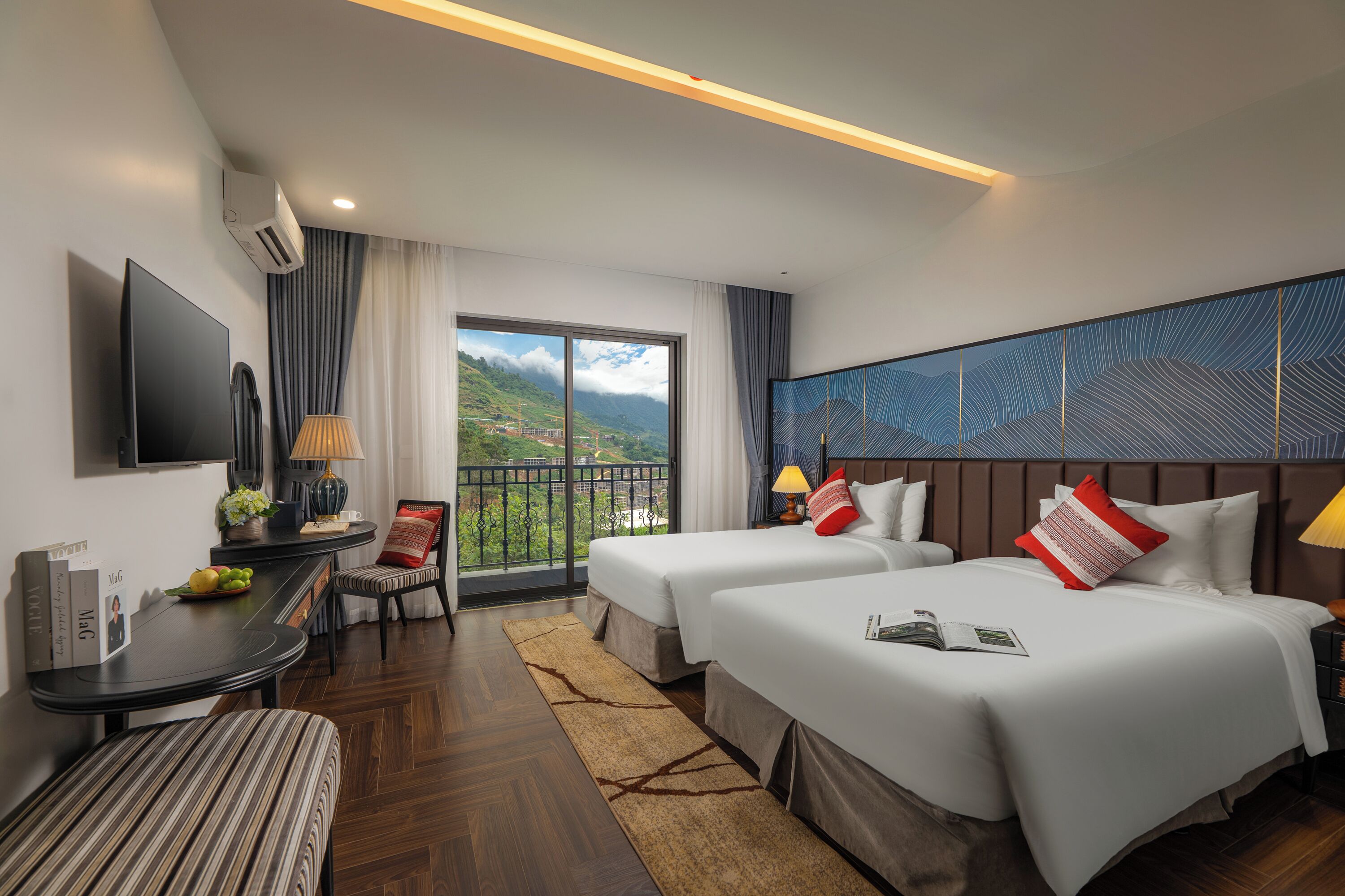 A hotel room with two beds, a TV, a desk with a lamp, a chair, and a view of the mountains.