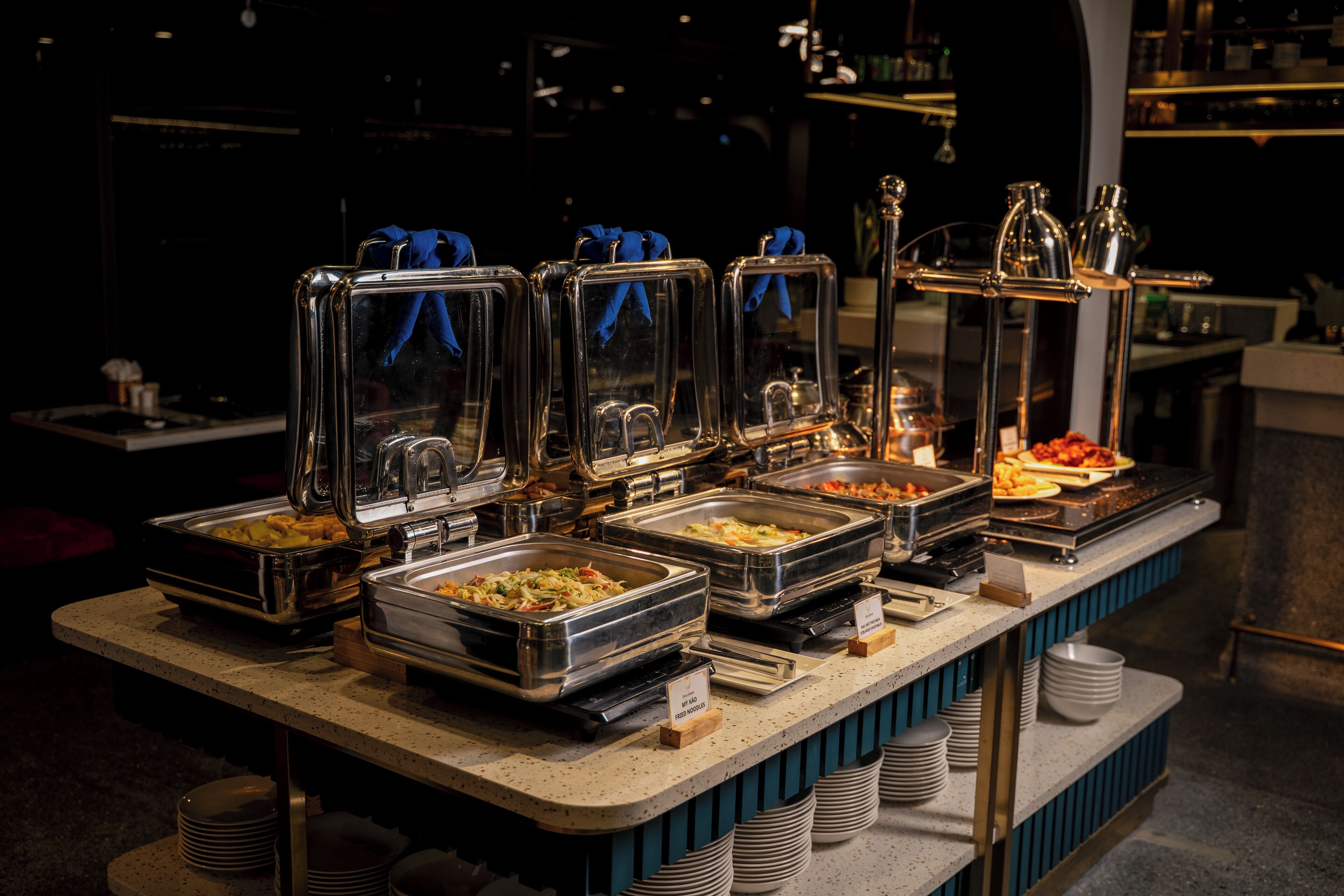 A buffet setup with various food stations, clear glass covers, and stacked white plates.