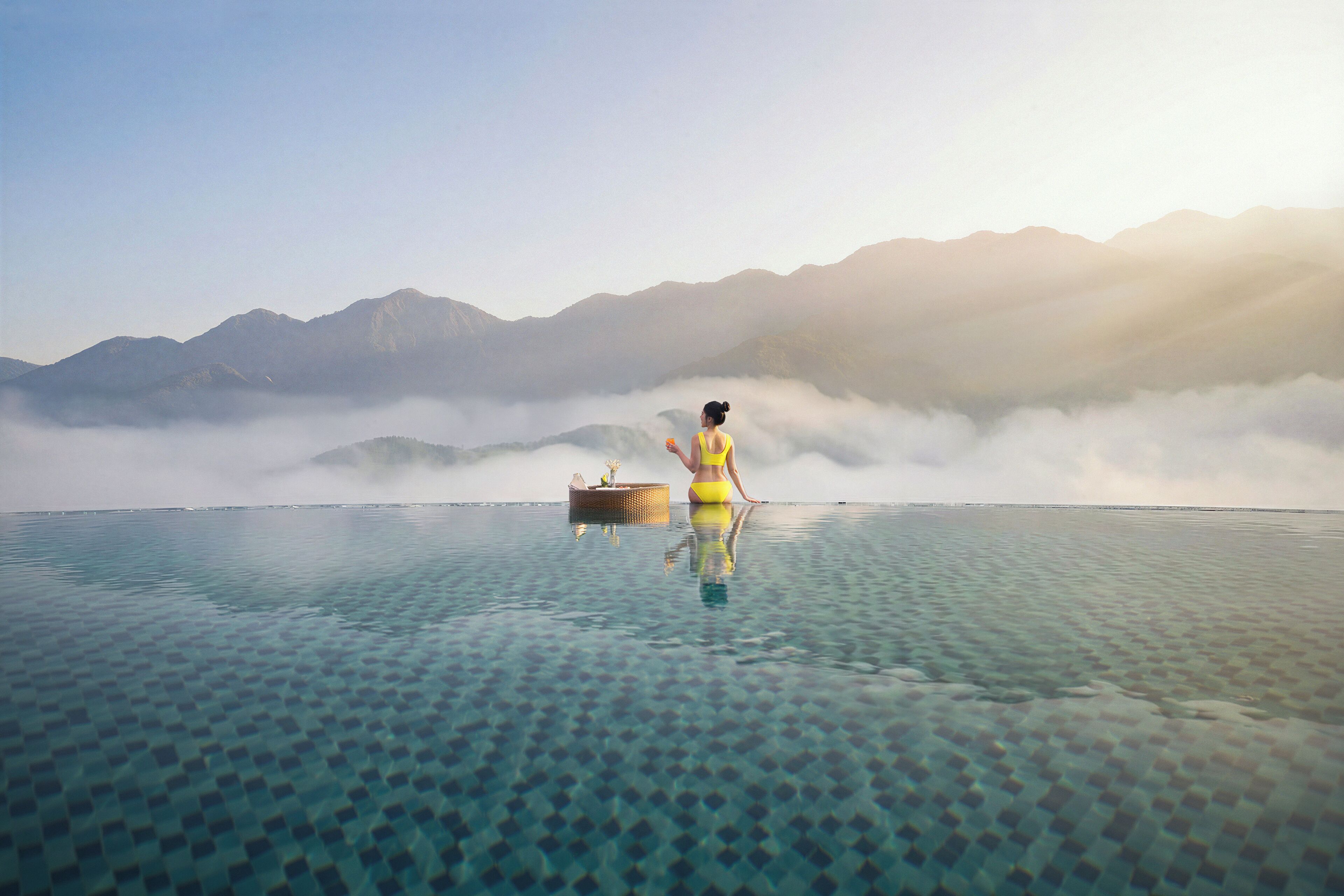 Una persona sentada en una plataforma flotante en una piscina cristalina con montañas al fondo.