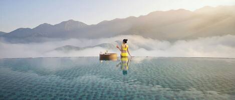 Una persona sentada en una plataforma flotante en una piscina cristalina con montañas al fondo.