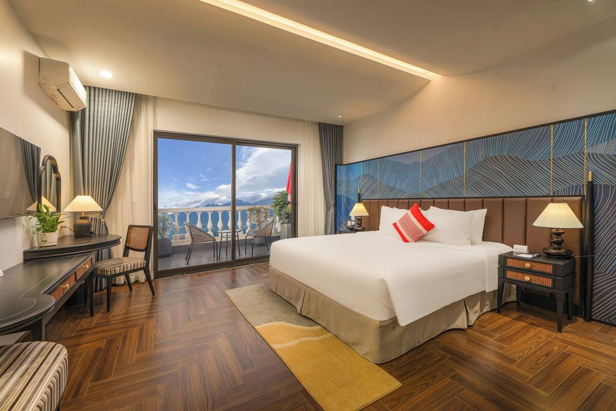 A hotel room with a large bed, a desk, a chair, a small table with a lamp, and a view of the outdoors.