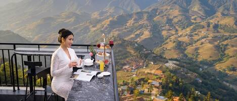 A woman sitting at a table with a view of mountains and a village below.