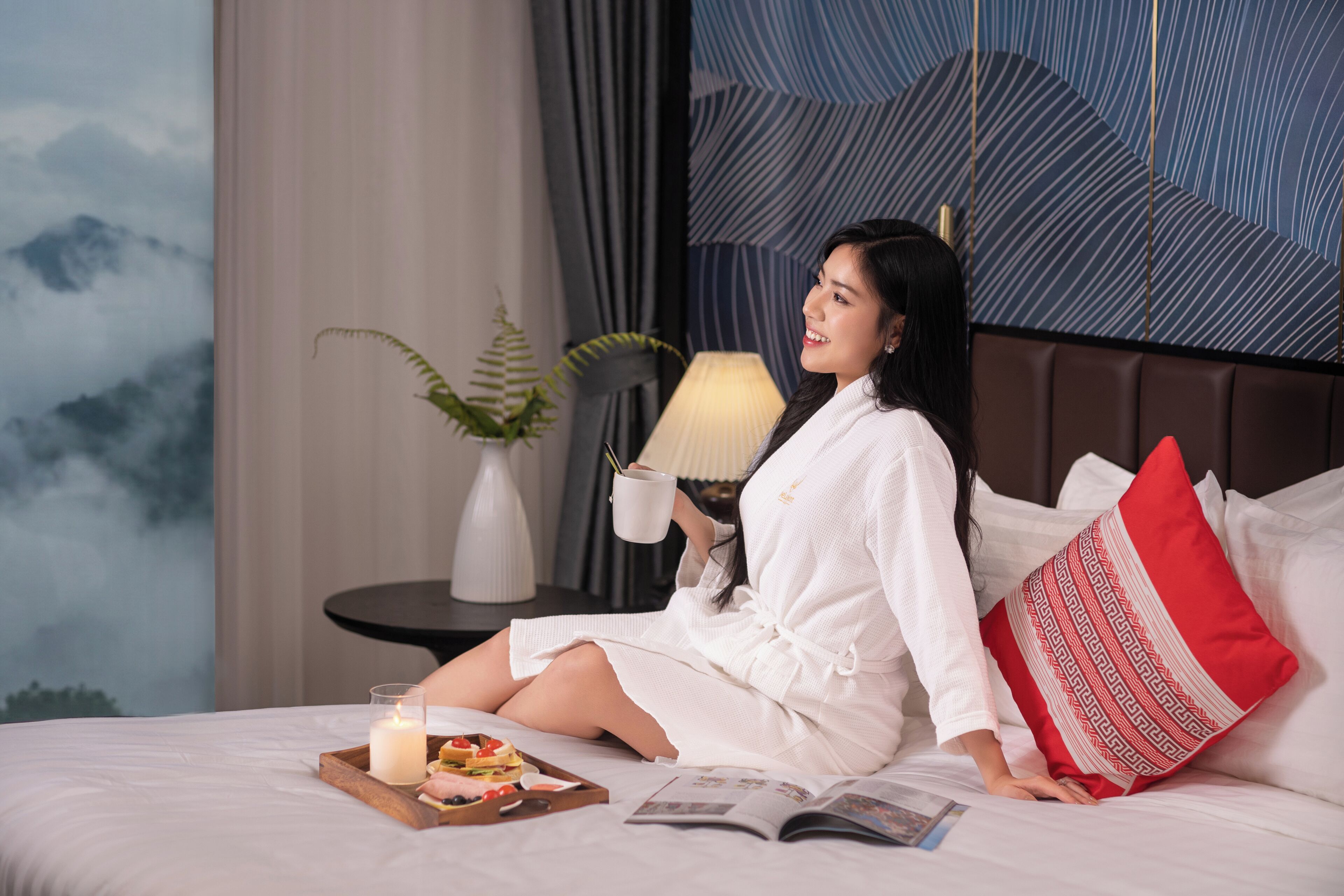 Una persona sentada en una cama, con un libro, una bandeja de comida y una vela.