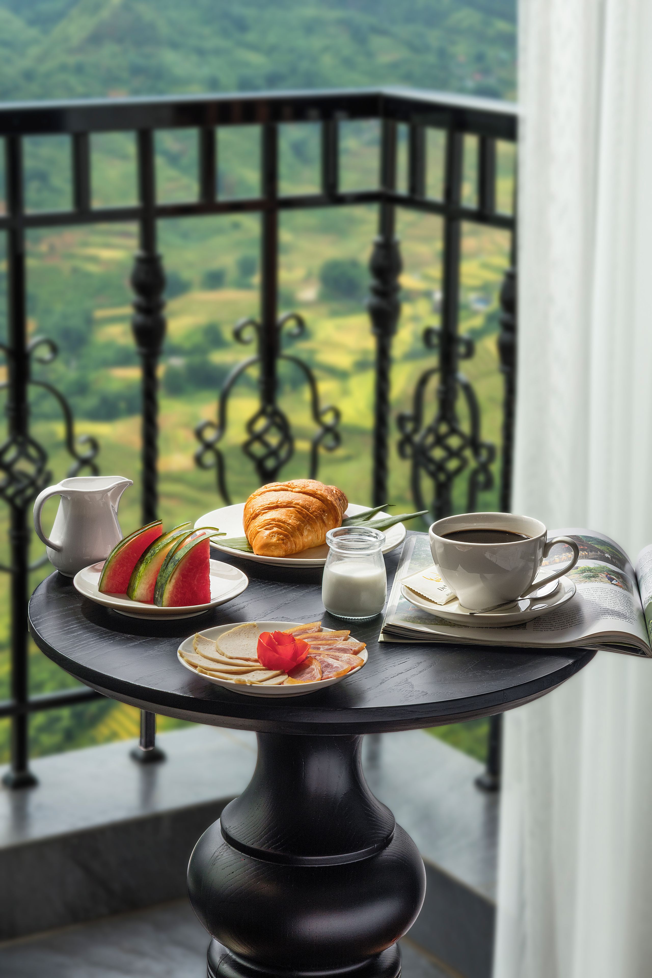 Una mesa con un croissant, una rebanada de sandía, una taza de café y una jarra de leche, frente a una vista panorámica.