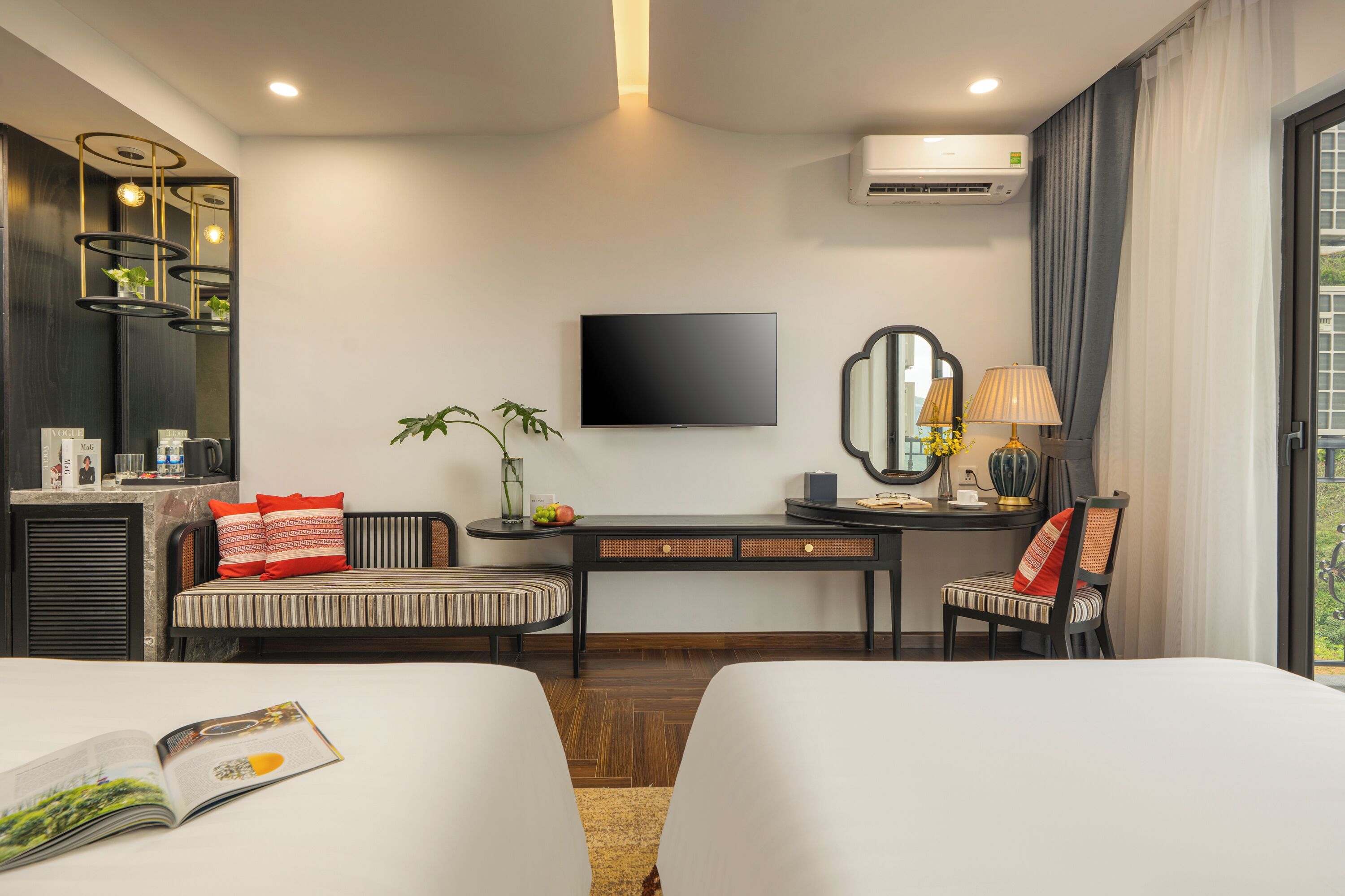 A hotel room with a bed, a desk with a TV, a chair, a plant, and a window with curtains.