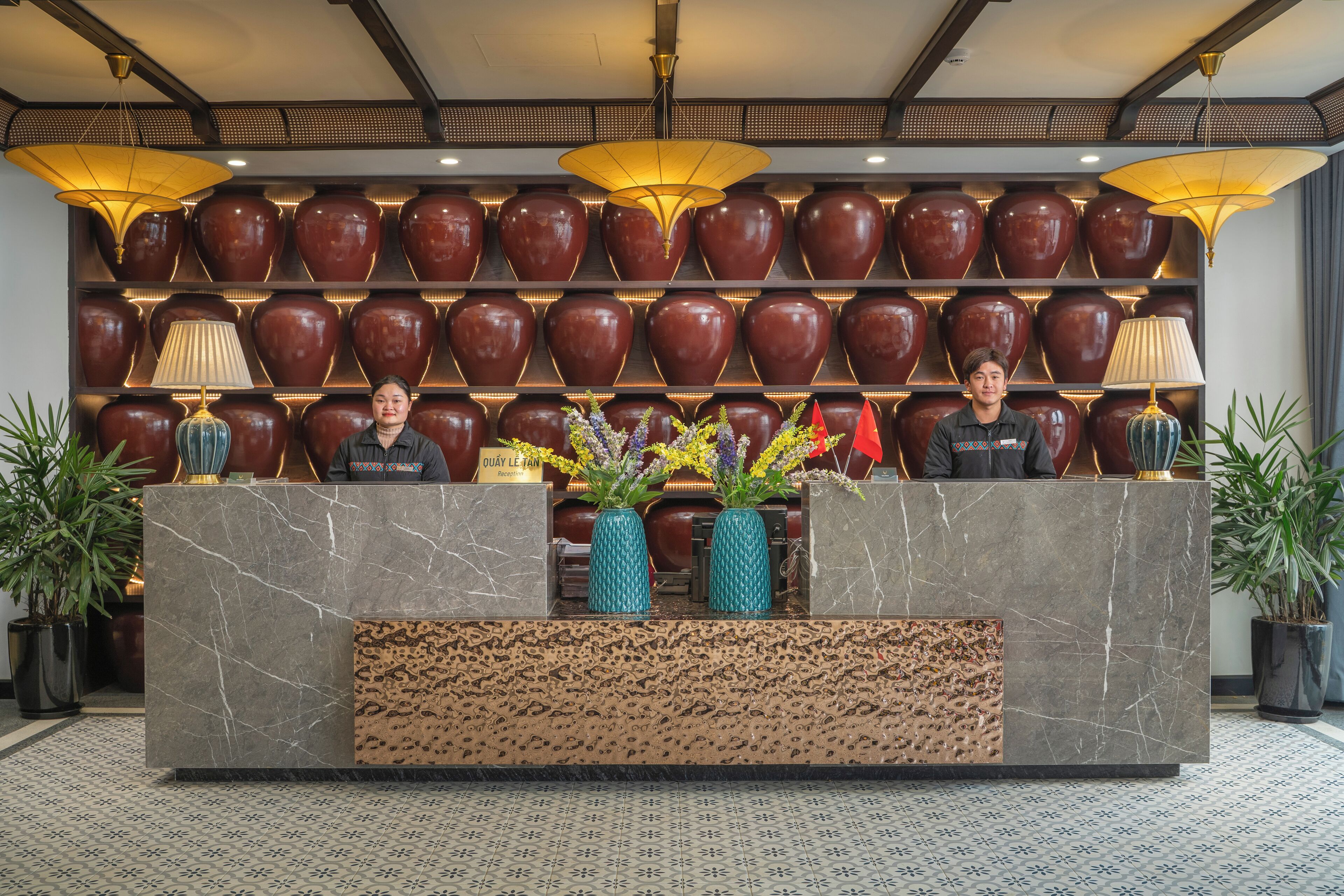 A reception desk with a marble top, two vases with flowers, and a wall of large vases behind.