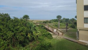 A wooden pathway leading to a beach, surrounded by lush greenery and a building with a beige exterior. - Family Beach Retreat with Ocean Views and Pools (Tybee Island)