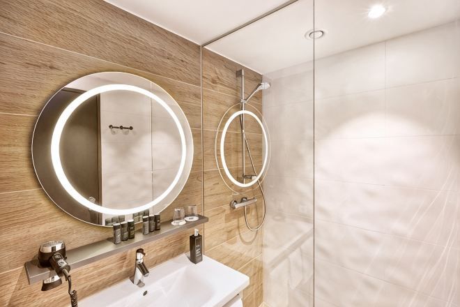 A modern bathroom with a wooden vanity, a round mirror with lights, a handheld showerhead, and a glass-enclosed shower.