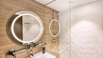 A modern bathroom with a wooden vanity, a round mirror with lights, a handheld showerhead, and a glass-enclosed shower.