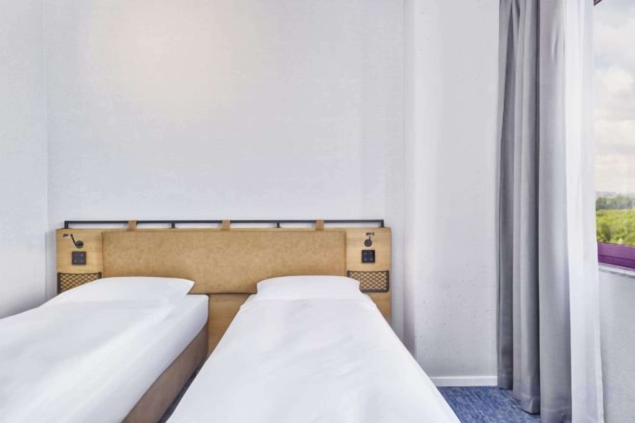 Habitación de hotel con dos camas individuales, cabecera, ventana con cortinas y vista a zonas verdes.