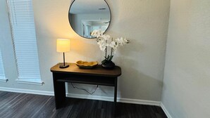 A small wooden console table with a round mirror, a floor lamp, and a vase with white flowers against a plain wall.