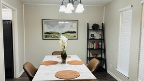 A dining room with a table set for four, surrounded by chairs, a chandelier, a bookshelf, and a painting on the wall.