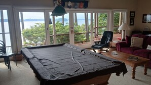Een kamer met een pooltafel, een paarse bank, een houten salontafel en uitzicht op zee door grote glazen deuren.