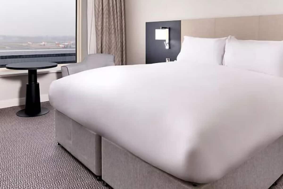 A hotel room with a large bed, a small table, a nightstand, and a view of the airport.