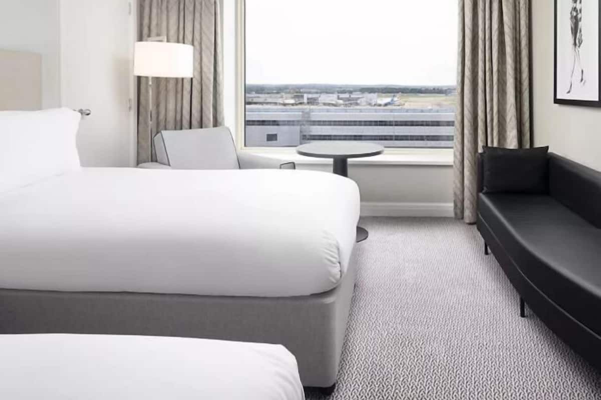 A hotel room with a large bed, a sofa, a small table, and a view of an airport.