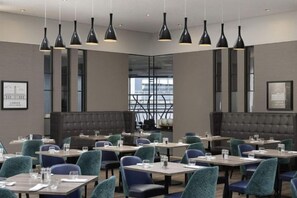 A modern dining area with multiple tables and chairs, pendant lighting, and a view through a large window. - Sofitel London Gatwick (Horley)