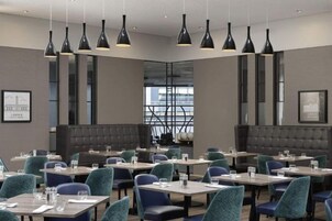 A modern dining area with multiple tables and chairs, pendant lighting, and a view through a large window.