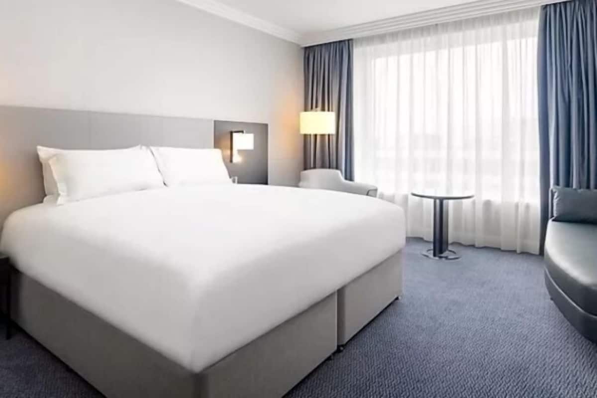 A hotel room with a large bed, a small round table, a chair, and a lamp.