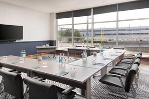 Un salón de conferencias con una mesa larga, sillas, un televisor de pantalla plana y ventanas amplias.