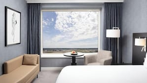 A hotel room with a large window, a bed, a sofa, a chair, a small table with a fruit bowl, and a lamp. - Sofitel London Gatwick (Horley)
