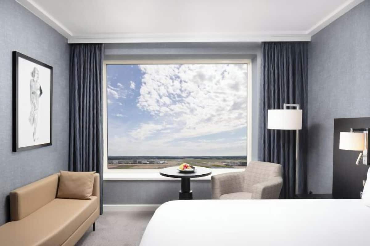A hotel room with a large window, a bed, a sofa, a chair, a small table with a fruit bowl, and a lamp.