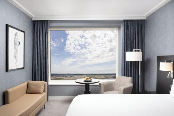 A hotel room with a large window, a bed, a sofa, a chair, a small table with a fruit bowl, and a lamp. - Sofitel London Gatwick (Horley)