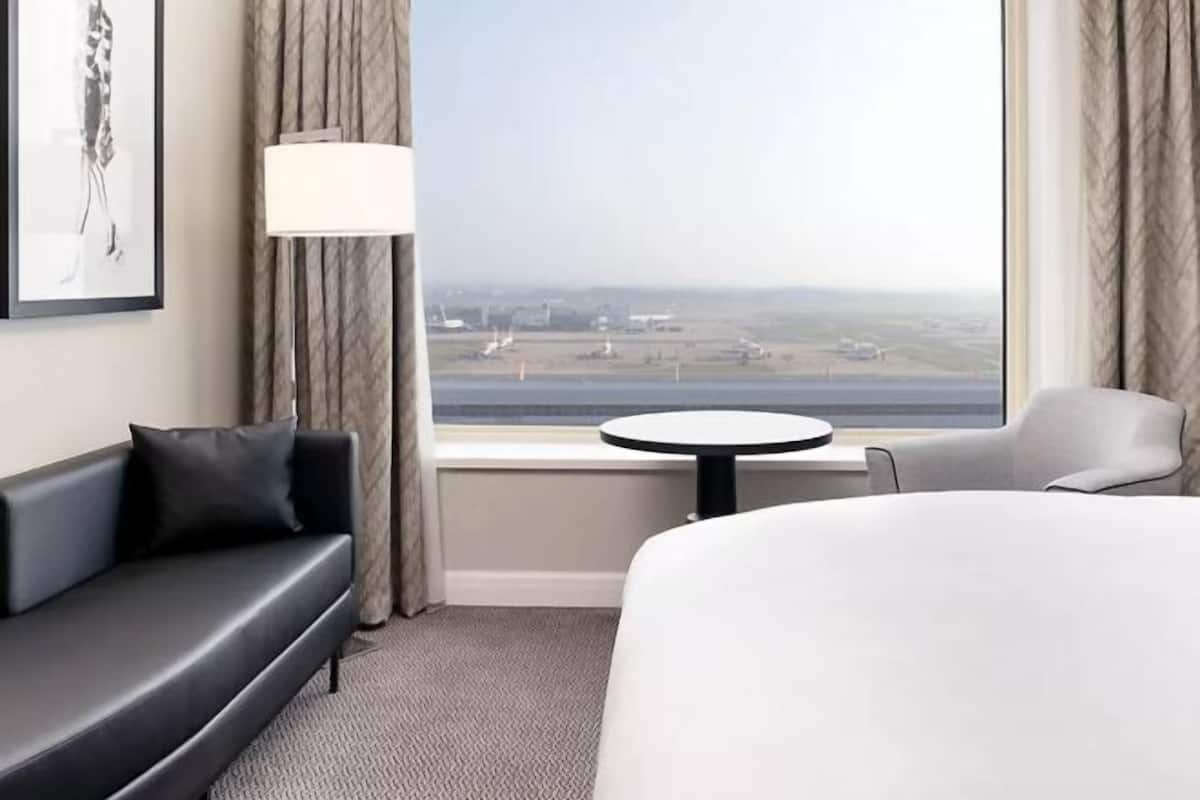 A hotel room with a bed, a sofa, a table, a lamp, and a view of an airport.