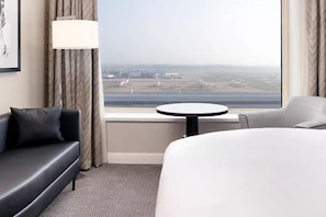 A hotel room with a large window offering a view of an airport. There is a bed, a sofa, and a small round table. - Sofitel London Gatwick (Horley)