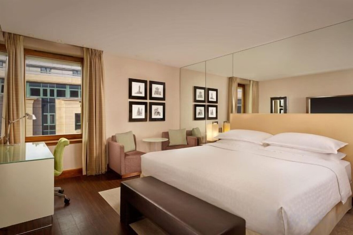 A hotel room with a large bed, a desk, a chair, a small table, and a bench.
