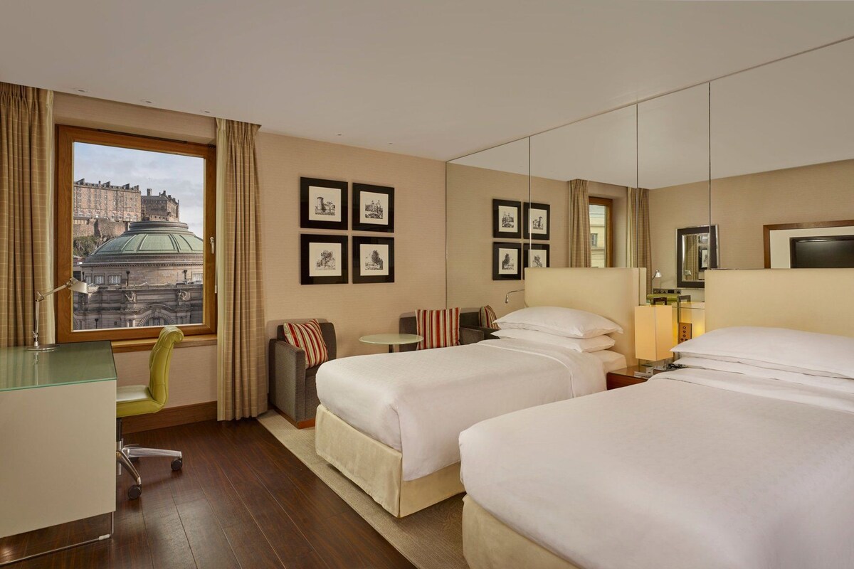 A hotel room with two beds, a desk, a chair, a small table, and a window with a cityscape view.
